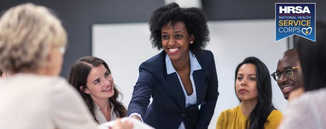 A standing, smiling woman leans forward while her colleagues look on.