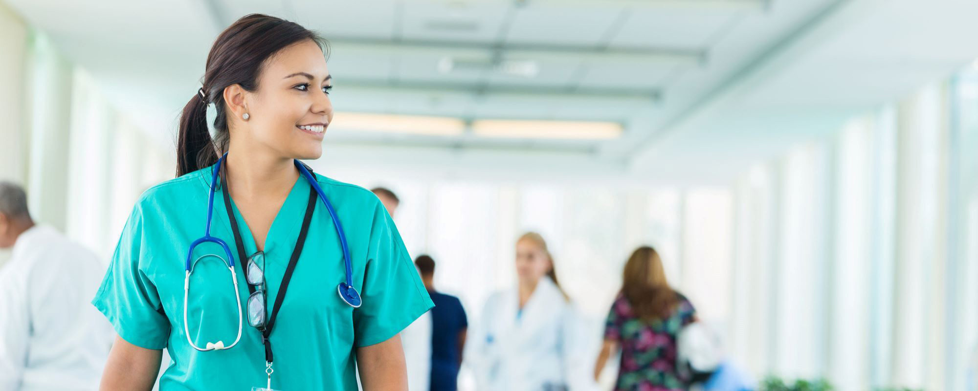 A health care professional wearing green scrubs and a stethoscope stands in a bright clinic hallway.